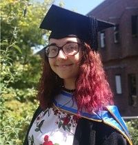 A woman in a graduation cap and gown, smiling in a garden. 